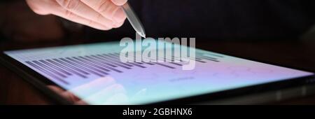 Male hand holds stylus over screen of tablet with commercial charts in dark Stock Photo