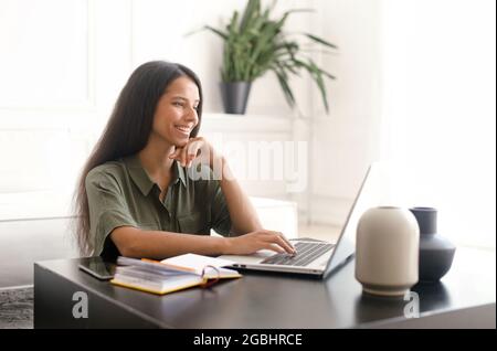 Cheerful indian young woman using laptop computer for remote work at home office. Smiling mixed-race businesswoman in casual wear holding video call, typing email, chatting online with colleagues Stock Photo