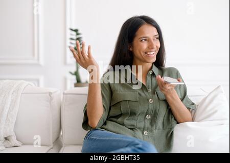 Charming young indian woman sits on the sofa at home, recording and sending voice message, mixed race female using voice recognition app on mobile phone, talking in the mic of the smartphone Stock Photo