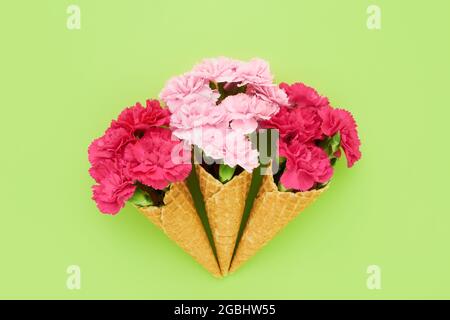 Three waffle ice cream cones with pink carnation flowers on a bright green background. Summer concept. Copy space for text, top view Stock Photo