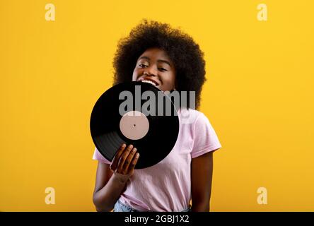 Joyful african american woman holding vinyl record, smiling and looking at camera on yellow background, copy space Stock Photo