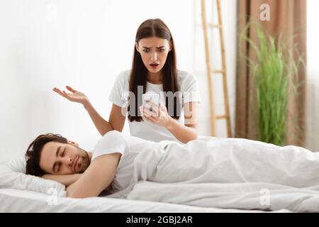 Girlfriend Holding Cheating Boyfriend's Phone Reading His Messages In Bedroom Stock Photo