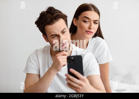 Jealous Girlfriend Peeking At Boyfriend's Cellphone Reading Messages In Bedroom Stock Photo