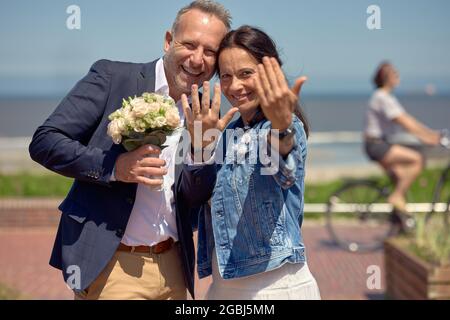 Happy romantic laughing newlywed couple showing off their rings as they pose together at the seaside with the husband holding the bridal bouquet in a Stock Photo