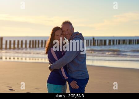Happy romantic middle-aged couple enjoying a loving cuddle on a deserted tropical beach at sunset standing in a close embrace smiling at the camera wi Stock Photo
