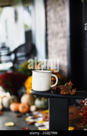 Hot steaming coffee sitting on rocking chair on front porch that has been decorated for autumn with heirloom white, orange, grey pumpkins, rain boots Stock Photo