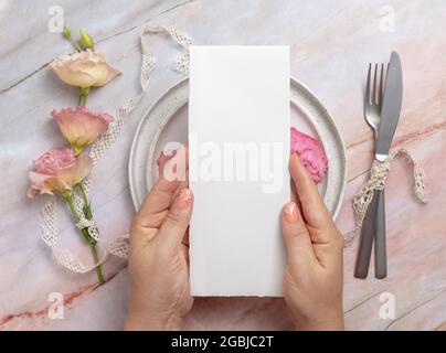 Hands holding wedding menu over a ceramic plate on a marble table decorated with flowers and ribbons. Mockup scene with blank paper menu card. Feminin Stock Photo