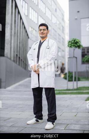 A young handsome Saudi Arabian male doctor smiling outside hospital. Full length portrait of male medical student in white coat, posing in front of modern university building or clinic Stock Photo