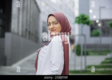 Middle Eastern healthcare worker. Closeup portrait of smiling confident muslim female nurse or doctor, wearing hijab, posing at camera outdoors in front of modern buildings Stock Photo