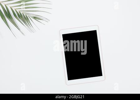 Tablet pc with blank screen next to palm leaf on white desk, flat lay. Stock Photo