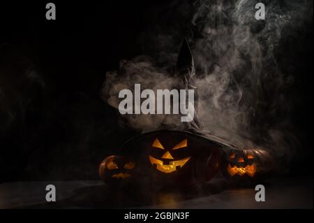 Three creepy Halloween grinning pumpkins glow in the dark among the fog. jack-o-lantern in a witch hat on a black background in smoke. Stock Photo