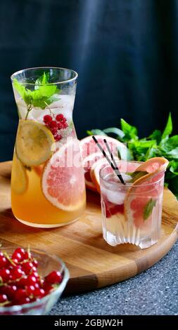 Grapefruit lemonade with mint, lemon, currants and ice in a decanter and a glass with black plastic tubes on a background of fruit. Cooling, refreshing, fruit, summer drink. Vertical close up Stock Photo
