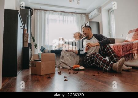 Moving day celebration. Yong couple Move Into New Home. Young happy couple, newlyweds family drinking wine and having fun in room with carton boxes in Stock Photo