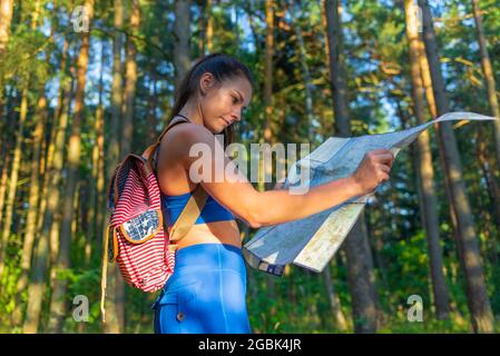 Young pretty hiker woman with backpack holding map in summer forest.female traveler checking destination on map.Young female hiker using map for orien Stock Photo