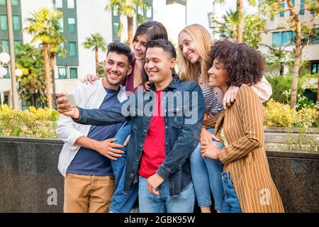 Group of five happy multicultural friends taking a selfie on a mobile phone on a city street laughing and smiling as they pose for the camera Stock Photo