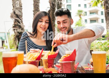Young couple eating crispy French Fries or potato chips at an outdoor table at a restaurant or pub in a city accompanied by glasses of cold beer Stock Photo