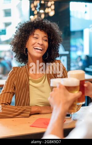 Happy vivacious laughing young Black woman toasting with a cold beer as she celebrates with a friend at a pub or restaurant in a city street Stock Photo