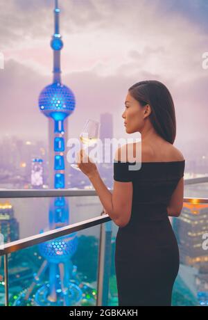 Luxury Shanghai lifestyle Asian woman drinking white wine in on China Travel. Elegant model in black dress at terrace view of the city, high end Stock Photo