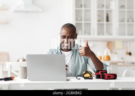 Review of construction instruments. Happy black handyman looking at laptop webcam and showing thumb up Stock Photo