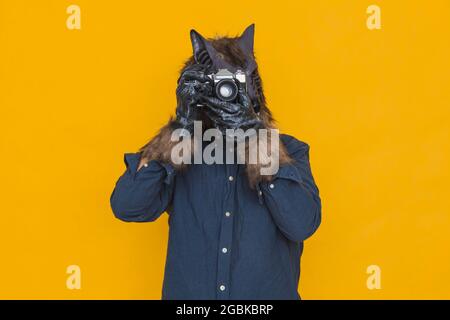 On a yellow background there is a werewolf dressed in a blue shirt is taking a photograph with an antique analog camera. Stock Photo