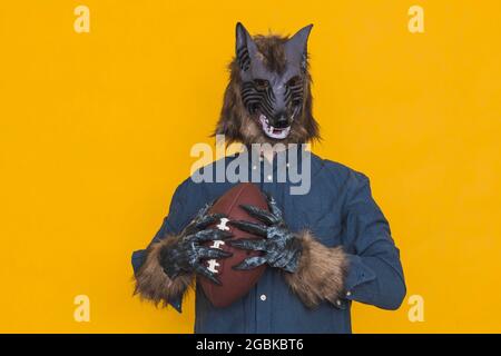On a yellow background there is a werewolf dressed in a blue shirt is holding a brown ball of american football. Stock Photo