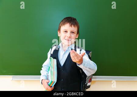 Funny schoolboy grimaces near the green school board in the classroom. Elementary school child with bag and books. Back to school. Stock Photo