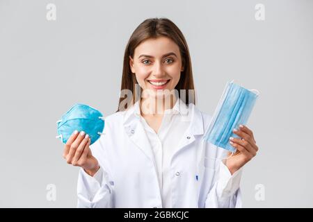 Covid-19, preventing virus, healthcare workers concept. Smiling attractive female doctor in white scrubs showing two variants of personal protective Stock Photo