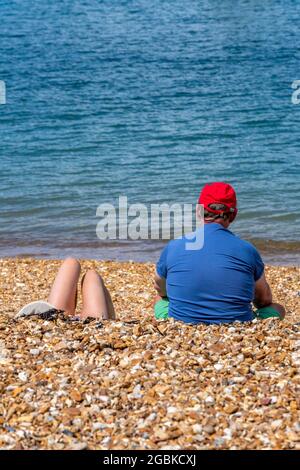 couple sunbathing sitting on a beach on the isle of wight, couple seated on pebble beach, middle aged couple, holiday on the beach, couple on beach. Stock Photo
