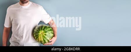 Young man keep whole watermelon in hands in white t-shirt on blue background. Mock up for design. Copy space. Stock Photo