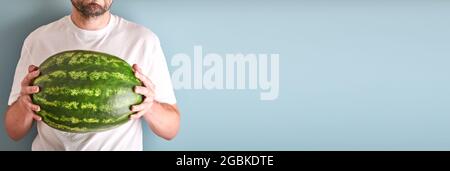 Young man keep whole watermelon in hands in white t-shirt on blue background. Mock up for design. Copy space. Stock Photo