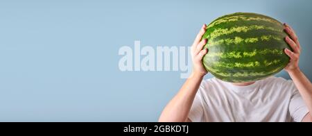 Young man keep whole watermelon in hands in white t-shirt on blue background. Mock up for design. Copy space. Stock Photo