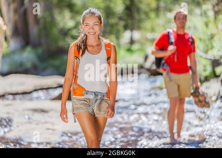 Hikers walking in forest crossing water stream holding boots hiking through Yosemite nature outdoors. Happy Asian girl and man in summer hike activity Stock Photo
