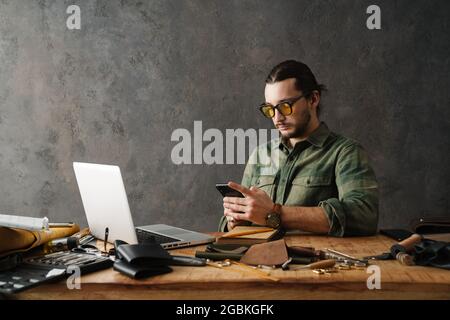 Bearded craftsman using laptop and smartphone while working with leather in workshop Stock Photo