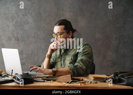 Bearded focused craftsman using laptop while working with leather in workshop Stock Photo