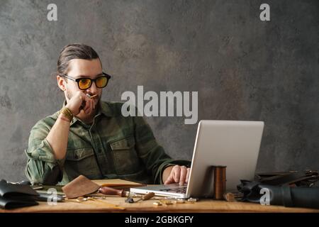 Bearded focused craftsman using laptop while working with leather in workshop Stock Photo