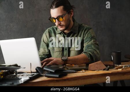 Bearded focused craftsman using laptop while working with leather in workshop Stock Photo