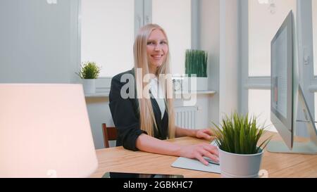 Beautiful young blond female sitting at workplace with computer and holding hand on mouse looking at camera and smiling Stock Photo