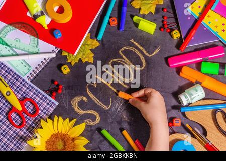 Children's hand writes the word "school" on a black chalkboard. School supplies as pencils, paints, scissors, colored notebooks, and others. Back to s Stock Photo