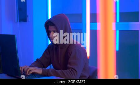 African hacker dressed in hoodie working on the computer in his secret place with warm neon lights Stock Photo