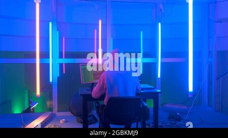 Back view of criminal hacker working in the evening on computer in big office with warm colored neon lights. Computer terrorism. Stock Photo