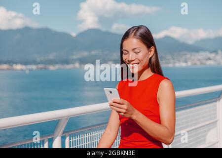 Asian young woman using mobile phone holding it in hand while walking commuting to work. Happy multiracial businesswoman smiling outside in Vancouver Stock Photo