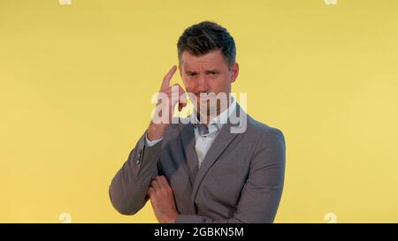 Close-up of man in suit putting his finger to the temple on yellow background. He is dissatisfied with something. Stock Photo