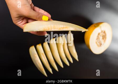 Female hand holding chunk of yellow melon in focus and rest of melon chopped on black background. Stock Photo
