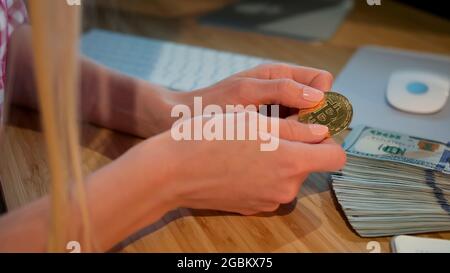 Crop view of hands of woman holding shiny metal bitcoin sitting at wooden desk with wad of cash nearby at night Stock Photo