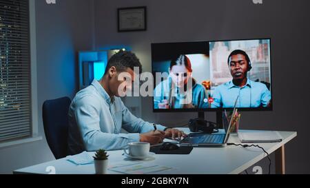 Black chief manager making video call on big monitor with colleagues on quarantine. They working home because of coronavirus pandemic. Stock Photo
