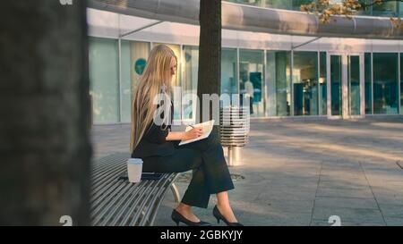 Side view of elegant businesswoman sitting on bench in patio and writing in notepad while working. Stock Photo