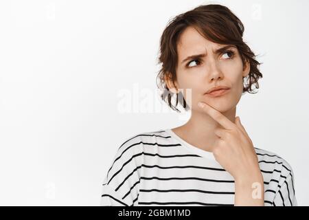 Hmm let me think. Thoughtful brunette woman frowning puzzled, looking aside and thinking, standing over white background Stock Photo