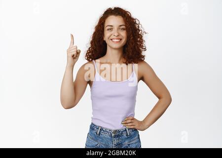 Attractive ginger girl, curly-haired woman pointing finger up, smiling and looking confident, showing advertisement, wearing summer tank top, white Stock Photo