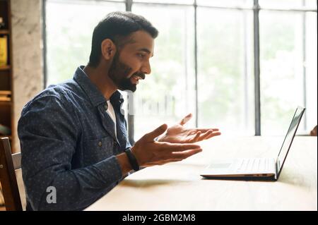 Confident smart indian freelancer guy working on laptop at home office, holding virtual meeting, multiracial eastern male employee using computer app for video call, involved virtual conference Stock Photo