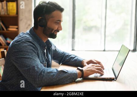 Young confident male Indian remote consultant wearing headset using laptop computer to virtual connection with customers or employees, customer service support manager talking at virtual meeting Stock Photo